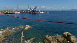 From a distance, the Costa Concordia surrounded by cranes, all floating on the calm waters.