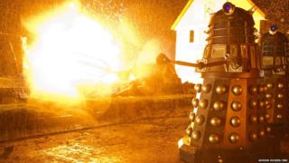 Two Daleks approach as flames erupt behind them.