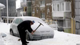 A man smashes ice with a pickaxe next to car covered with ice