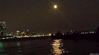 The orange moon sits above an illuminated skyline, next to the ocean, and reflects in the water.