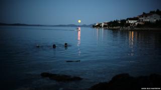 The yellow moon is hanging above the sea, being reflected in the water, near to land.