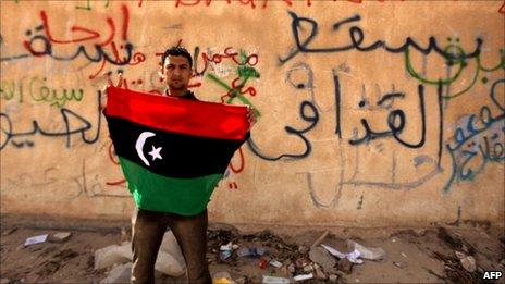 A Libyan anti-government protester holds his old national flag in front of a wall covered with graffiti against Libyan leader Muammar Gaddafi in the eastern city of Tobruk on February 24, 2011