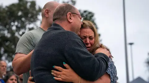 Matilda's father, dressed in a grey pullover, embraces her weeping mother, dressed in a blue and white t shirt