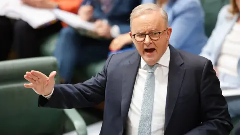 A middle aged man with thinning grey hair and glasses, wearing a suit and tie, gestures as he addresses parliament