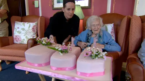 Centenaria Hilda Leggett sitting in an amrchair in front of a pink birthday cake in the shape of the numbers 110