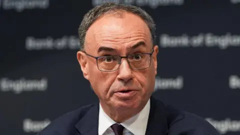 Andrew Bailey, wearing a dark suit, raises his eyebrows while seated in front of a dark, "Bank of England" branded background.