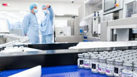 two people in blue medical suits, hairnets and masks stand behind a conveyor belt that is covered with vials of clear medicines