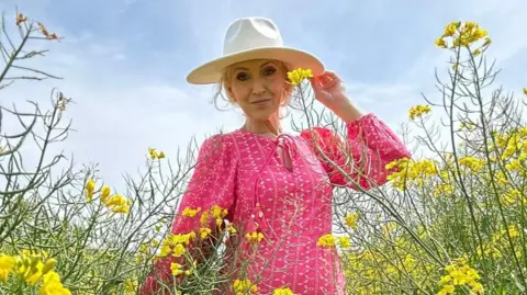 A woman wearing a pink dress and a hat. She is pictured in a field with yellow flowers around her.