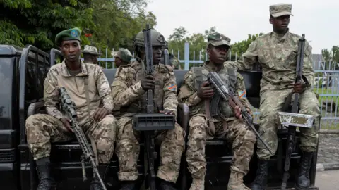 Four M23 fighters wearing military uniforms and holding guns sitting on a truck