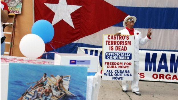 An anti-Castro protester flashes a victory sign as he stands in front of a Cuban flag (rear) and near a booth promoting human rights in Cuba (bottom left) during a small anti-Castro rally in Miami, Florida 08 November 2006.