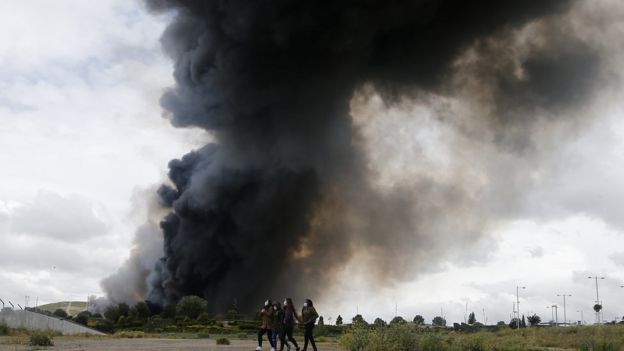 four women in face masks walk in front of a pillar of thick black smoke, May 13 2016