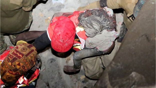 A young girl with ash-grey face, legs and hair is carried by a rescuer