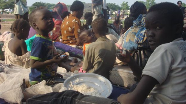 two children, one about 5 and the other about 12 years old, eating from a plate of rice while sitting on blankets surrounded by other people, 11 July