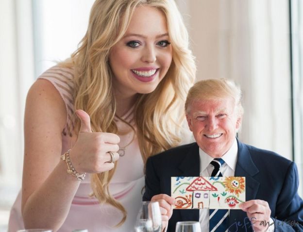 A small Trump holding up a child's picture, with his daughter Tiffany doing a thumbs up over his shoulder