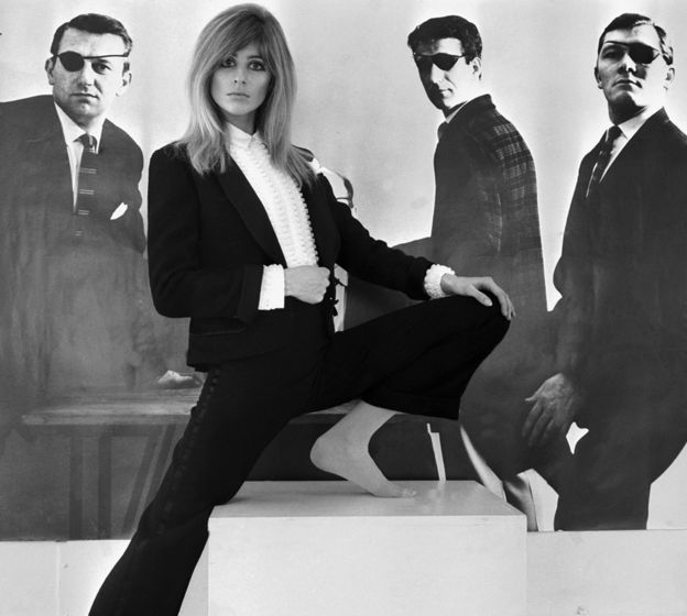 British model Jill Kennington posing in front of a picture of suited men wearing eyepatches, 1967
