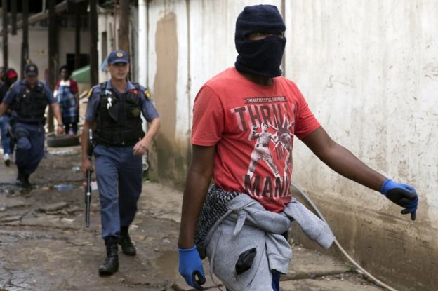 SA national wearing balaclava and carrying a rock walks ahead of an armed policeman