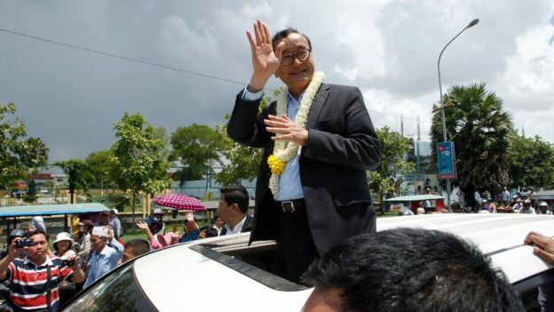 Sam Rainsy stands on the seat of a car, waving to crowds in Phnom Penh and wearing a flower garland around his neck