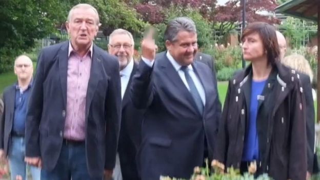 Sigmar Gabriel, surrounded by people, flicking his middle finger in the direction of the camera