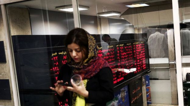 An Iranian woman uses a mobile phone next to a stock market activity board at the stock exchange in the capital Tehran