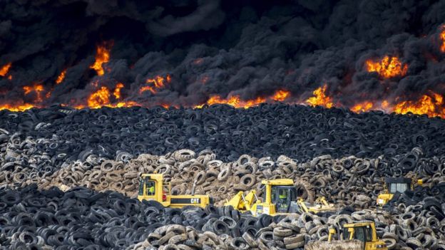 piles of intact tyres behind industrial diggers, with flames and smoke in the background, May 13 2016