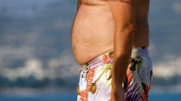 Homem com muita gordura abdominal de bermuda na praia