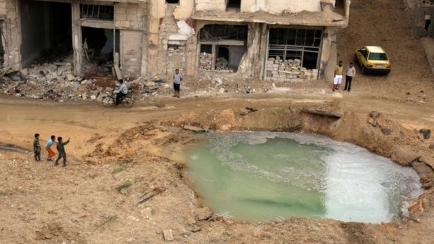 People inspect a hole in the ground filled with water in a damaged site after airstrikes on the rebel held Tariq al-Bab neighbourhood of Aleppo