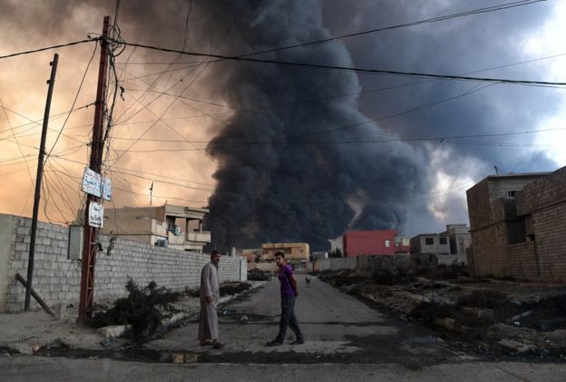 Hombres en la calle frente a un incendio provocado por la retirada de EI en Qayyarah, Irak