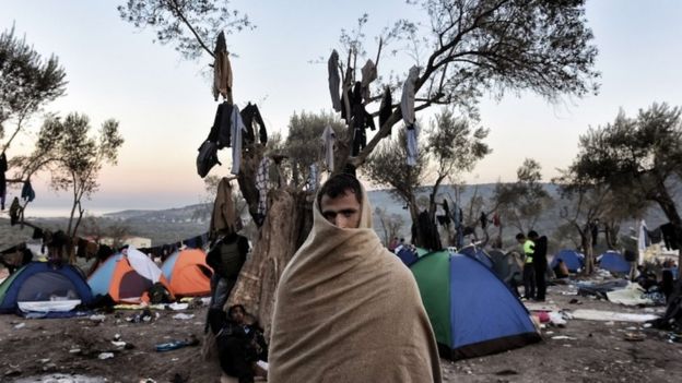 A man with a blanket wrapped around his head and shoulders stands near tents where refugees and migrants live in a field outside the Moria registration center on the Greek island of Lesbos on November 11, 2015