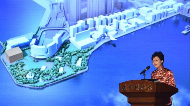Carrie Lam introducing the museum at a podium, with a screen showing an image of the proposed building behind