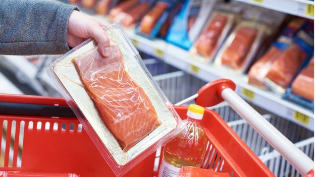 Imagem mostra mulher pondo um pacote de salmão em um carrinho de supermercado