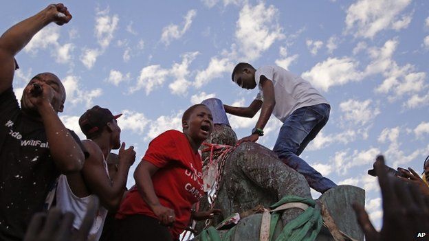 Students shout out and place a bucket on the head as they climb on top of the decades old bronze statue of British colonialist Cecil John Rhodes, as the statue is removed from the campus at the Cape Town University