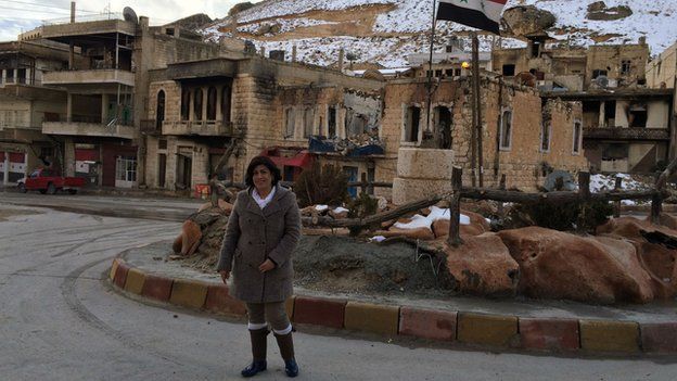 Antoinette Nasrullah in Maaloula, standing in front of wrecked buildings (including her own)