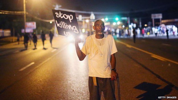 Older man holding a sign that says "stop killing us"