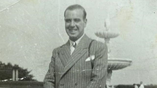 An old black and white picture of a man in a suit with a camera slung over his shoulder