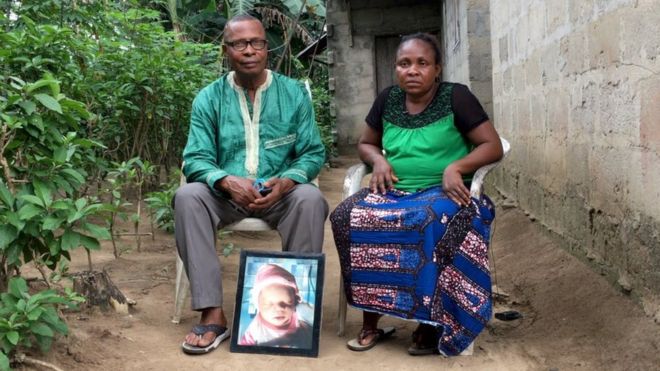 Barinaadaa Saturday e Chief Bira Saturday sentados do lado de fora da casa, com um retrato do filho recém-nascido morto.
