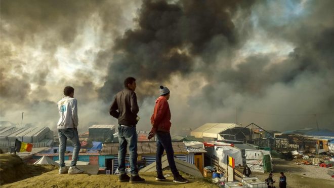 Migrants stand on a hill overlooking the "Jungle" migrant camp in Calais, northern France, as smoke rises on October 26, 2016