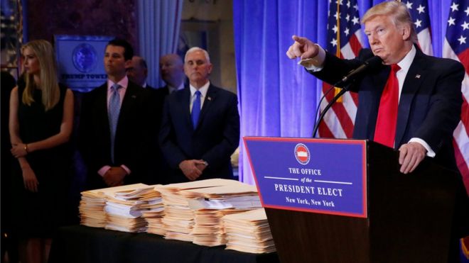 Donald Trump points at the press while standing next to a table covered in documents at a news conference