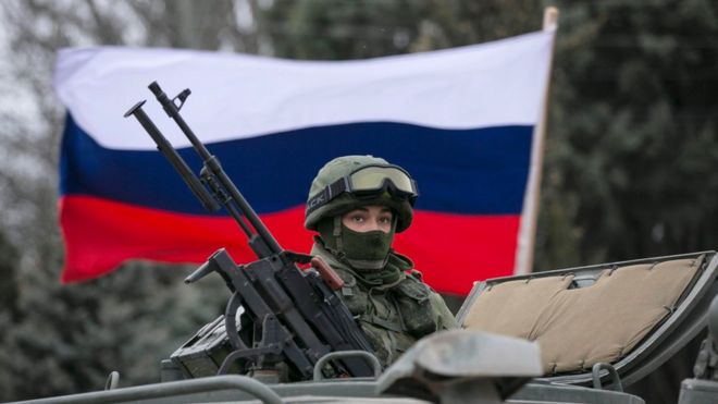 A pro-Russian man (not seen) holds a Russian flag behind an armed servicemen on top of a Russian army vehicle outside a Ukrainian border guard post in the Crimean town of Balaclava March 1, 2014.