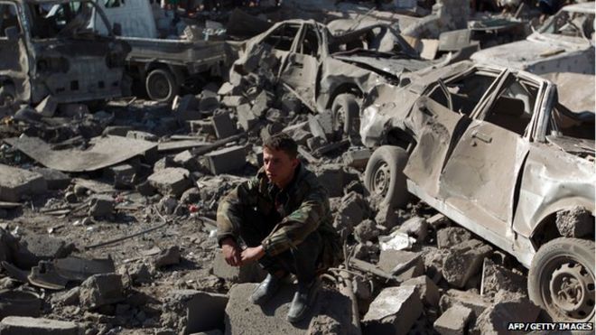 A Yemeni man wearing a military fatigue sits above debris at the site of a Saudi air strike against Houthi rebels near Sanaa Airport 26 March 2015
