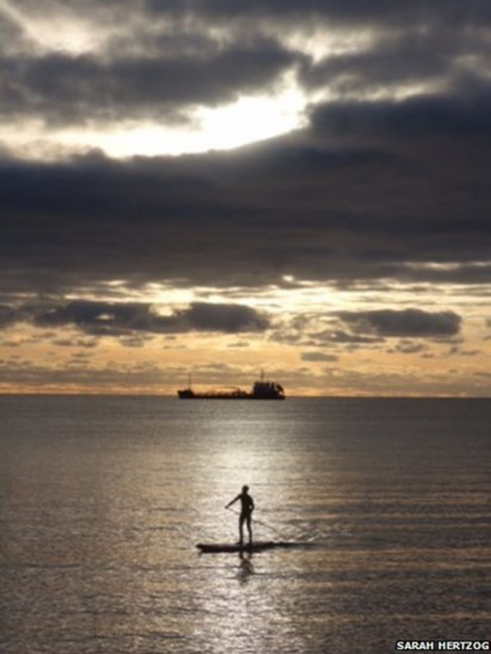 A figure on a paddle board in the sea is silhouetted in the sunrise