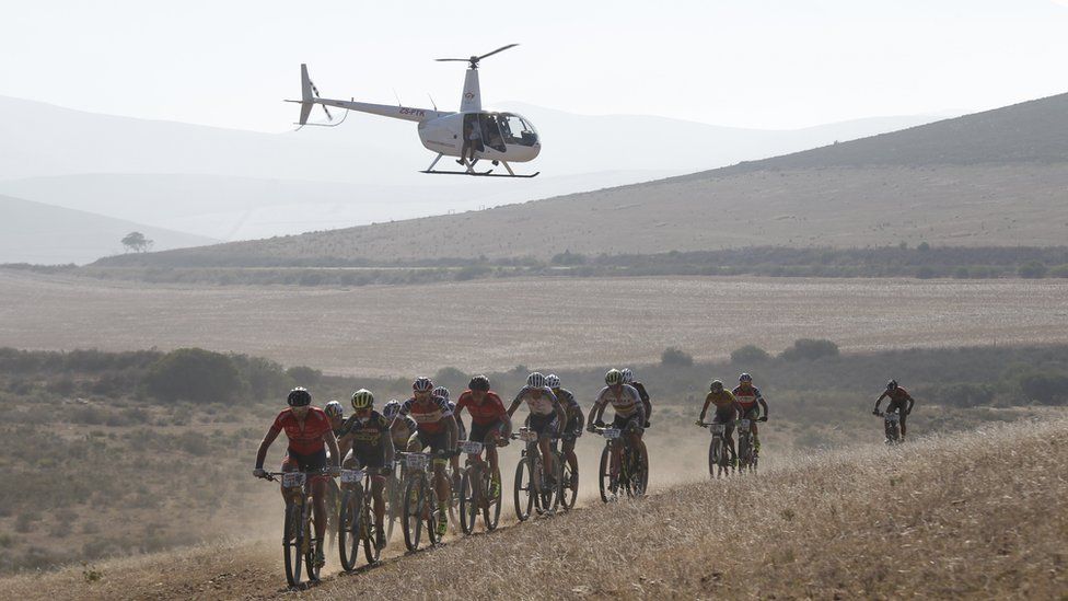 Professional riders taking part in The professional peleton race cycle on a track in Greyton, South Africa, while a helicopter hovers overheads.