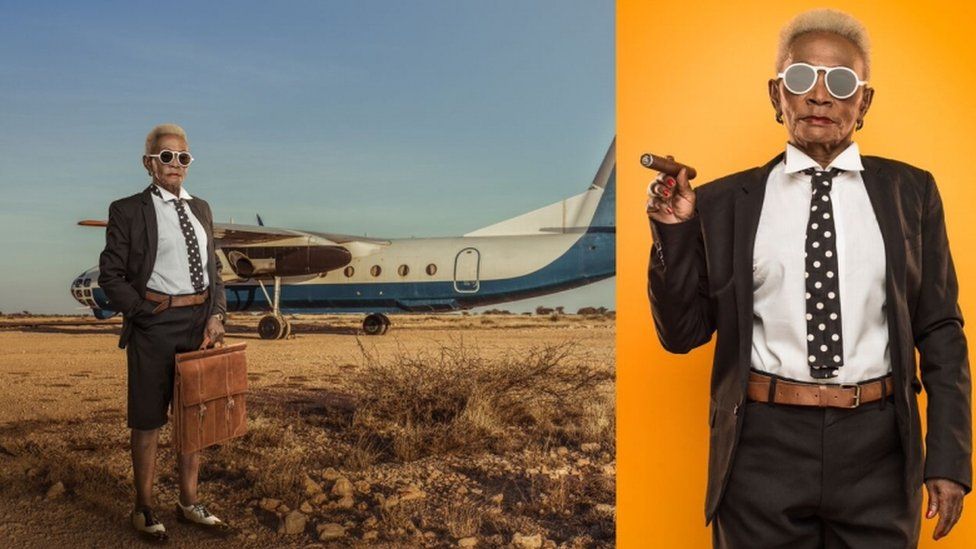 A woman with grey hair dressed as a businessman with tie, jacket, cigar and brief case - standing by a plane - in a work by photographer Osborne Macharia