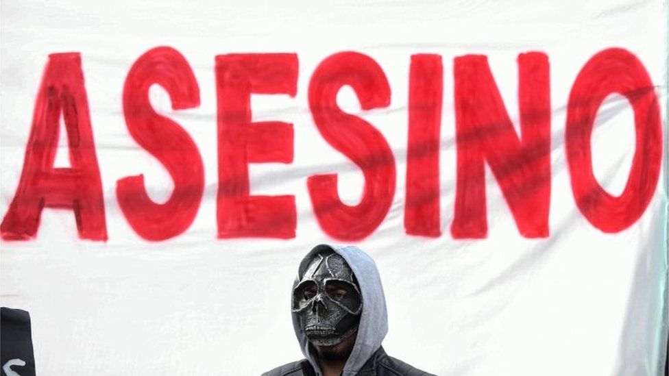 A man with a mask stands before a placard reading "murderer"