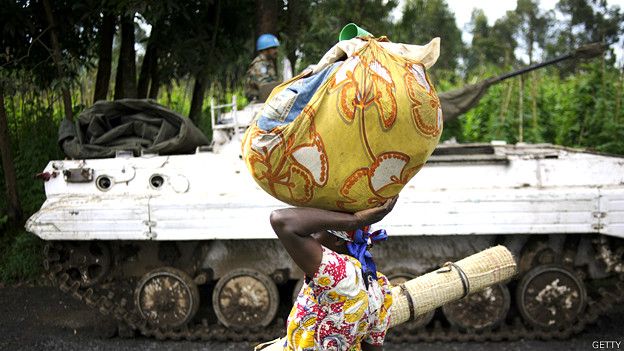Una mujer en África camina con un bulto de ropas en su cabeza frente a un tanque conducido por soldados de la ONU