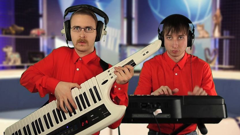 Brett Domino playing Key-tar in a red shirt next to his sidekick steve playing the keyboard super imposed into the Blue Peter studio.