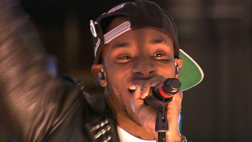 Rapper Angel holding a mocrophone to his mouth and singing. Wearing a black baseball cap backwards.