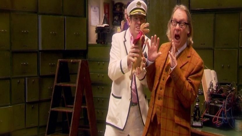 Vic Reeves and Captain Lengthwidth holding a T-Rex in a cape.