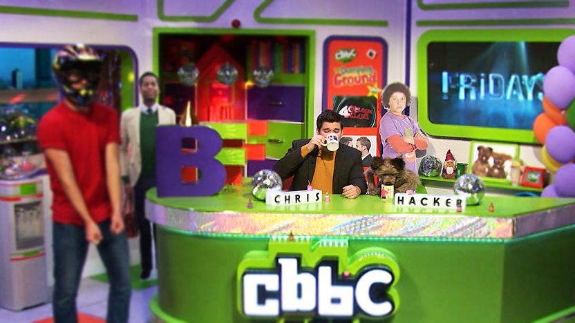     Chris Johnson and Hacker T Dog in the CBBC Office drinking a cup of tea while a man in a motorcycle helmet is stood to the left of the desk.