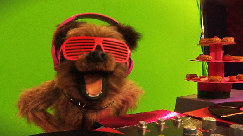 Hacker T Dog wearing rave glasses and headphones in front of a green screen.
