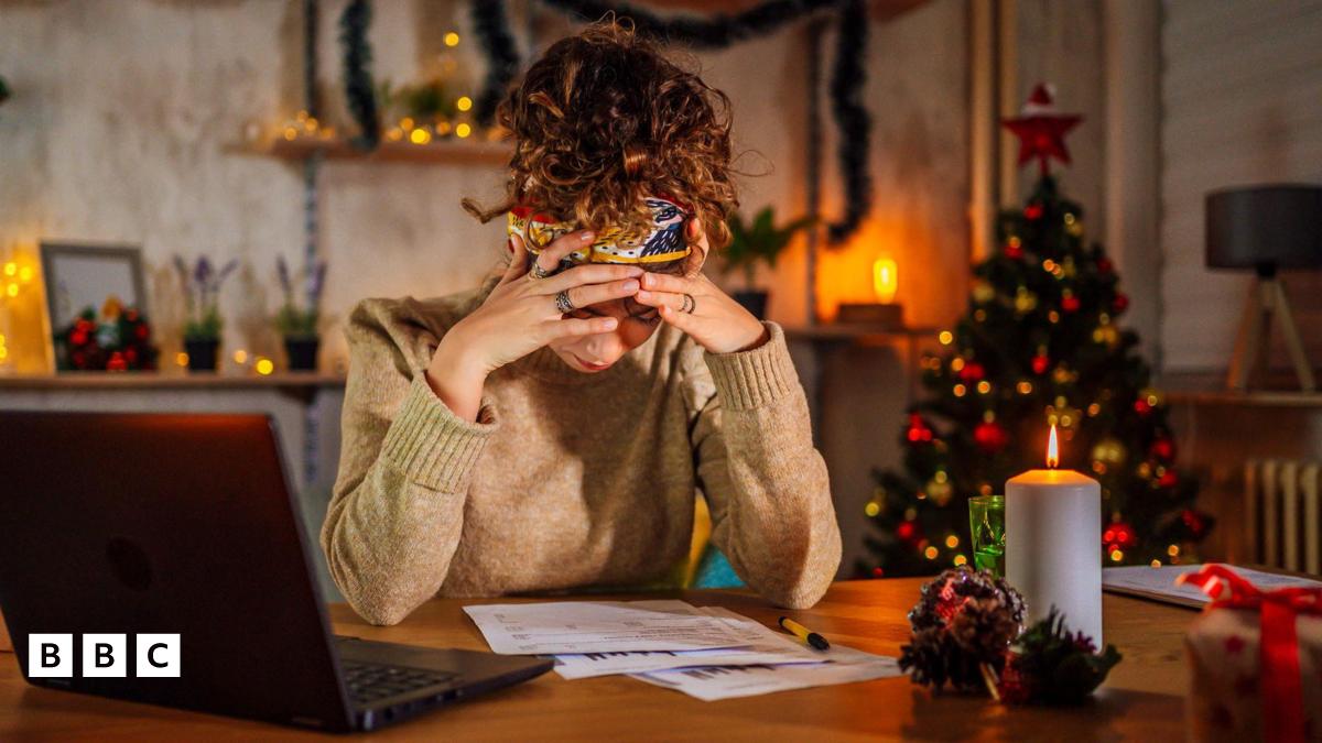 woman in beige jumper in front of a computer with her head in her hands. Xmas tree in the background